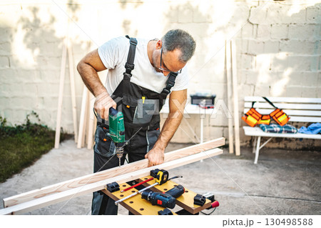 Mature male carpenter using electric drill on a wooden plank outdoors Mature male carpenter using electric drill on a wooden plank outdoors 130498588