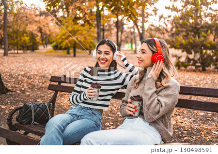 Two women sitting on a park bench with headphones and drinking coffee Two women sitting on a park bench with headphones and drinking coffee 130498591