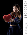 Smiling young woman in a navy blue outfit holding out a fluffy red heart pillow against a black background, symbolizing love 130499894