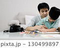 Loving father sits closely with his young son, guiding him as he draws with colored pencils at a table, creating togetherness 130499897
