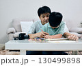 Father sits closely with his young son at a table, guiding him as he works on a drawing with colored pencils at home 130499898