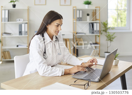 Young cheerful woman doctor sitting at the desk on workplace and working in medical clinic. Young cheerful woman doctor sitting at the desk on workplace and working in medical clinic. 130503776