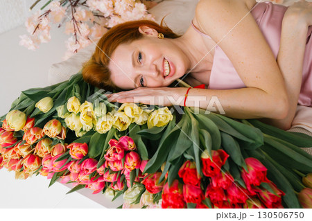 Caucasian female lying with colorful tulips and cherry blossoms, smiling in pink dress 130506750