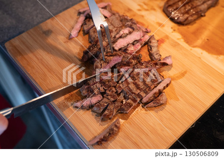 Grilled steak slices on wooden cutting board being cut with knife and fork Grilled steak slices on wooden cutting board being cut with knife and fork 130506809
