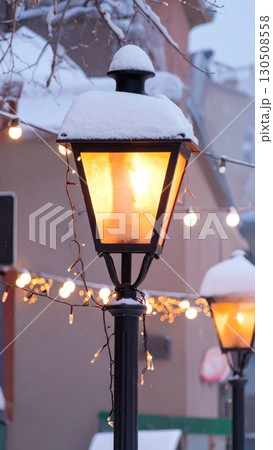 Snow Covered Black Street Lamp Glowing Warmly at Dusk in Winter City Landscape 130508558