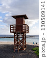 A weathered wooden lifeguard tower stands tall on a sandy beach under a partly cloudy sky, watching over the calm waters. 130509141