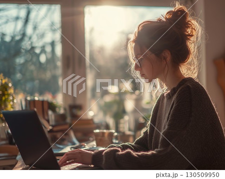 A young woman with glasses focuses intently typing on her laptop in warm golden light 130509950