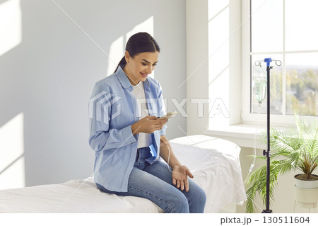 Young smiling woman using smartphone while receiving IV drip infusion and vitamin therapy. 130511604