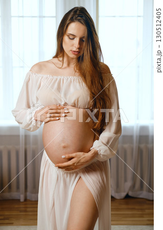 Pregnant woman in soft attire gently cradling her belly in a bright indoor setting Pregnant woman in soft attire gently cradling her belly in a bright indoor setting 130512405