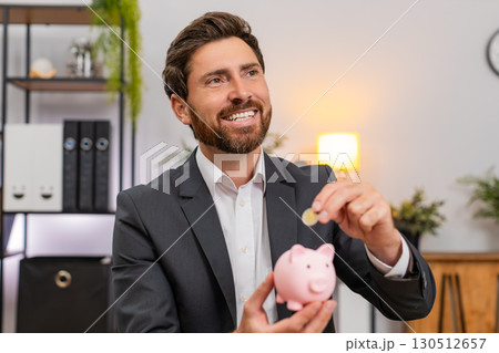 Middle-aged businessman puts coins into piggy bank plans future savings and investments with smile 130512657