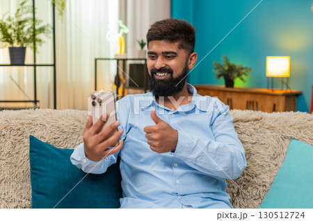 Indian man scrolling smartphone news feed liking post thumbs up happy reaction social media indoors Indian man scrolling smartphone news feed liking post thumbs up happy reaction social media indoors 130512724