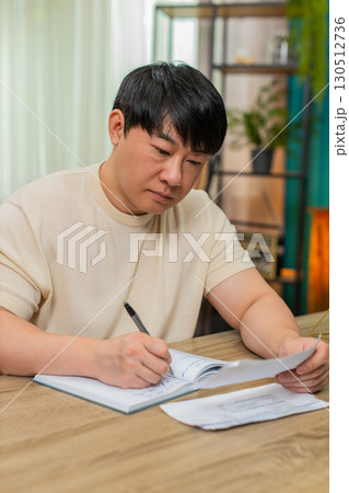Asian man studying utility bills, frowning, adding notes in notebook, focused financial calculations Asian man studying utility bills, frowning, adding notes in notebook, focused financial calculations 130512736