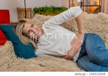Adult woman holding stomach at home, expressing abdominal pain and discomfort while lying on sofa Adult woman holding stomach at home, expressing abdominal pain and discomfort while lying on sofa 130512838
