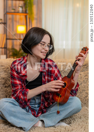 Young woman playing ukulele at home, focused on learning chord and smiling with satisfaction and joy 130512840