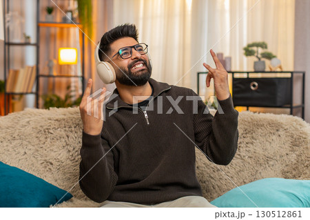Indian man smiling listens to energetic music in wireless headphones, shows rock-n-roll gesture 130512861
