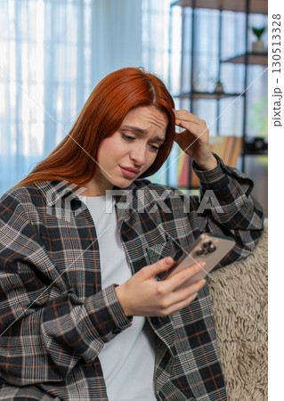 Woman sitting on sofa holding smartphone looking disappointed at screen after bad message or news 130513328