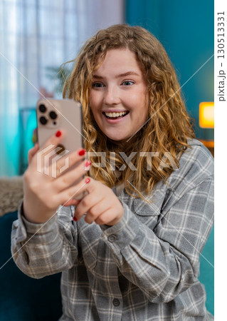 Young woman sitting on sofa smiling with joy reacting to long awaited message on smartphone happily 130513331