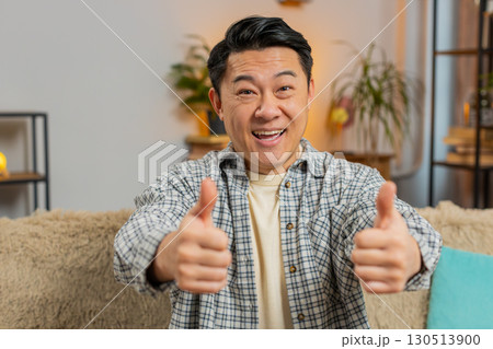 Portrait of excited smiling Chinese young man showing double thumbs up sitting on sofa couch at home Portrait of excited smiling Chinese young man showing double thumbs up sitting on sofa couch at home 130513900