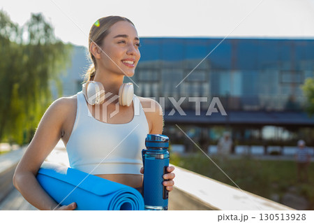 Happy young sportswoman jogger with water bottle and rolled mat walking in city park on sunny day 130513928