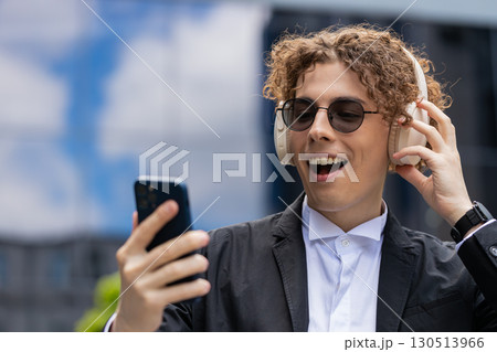 Happy joyful businessman listens to music on wireless headphones while walking on street near office Happy joyful businessman listens to music on wireless headphones while walking on street near office 130513966