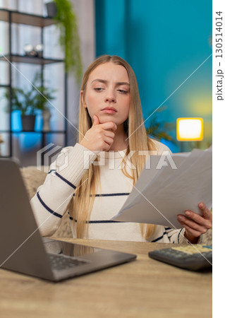 Caucasian woman managing tax declaration at home on sofa table, holding documents, calculator laptop 130514014