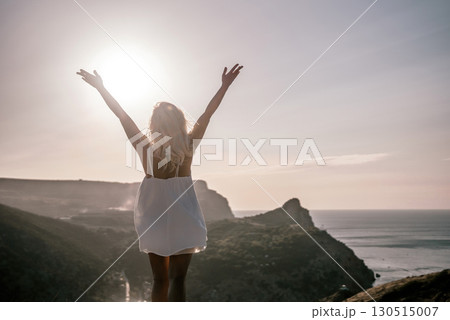 A woman is standing on a hill overlooking a body of water. She is wearing a white dress and she is happy. 130515007