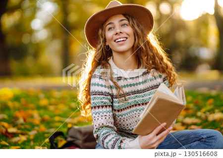 Young woman in hat sits on a plaid in autumn park and reads a book. Concept of nature, education. 130516608