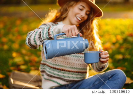 Beautiful woman in stylish clothes enjoys hot drink in hat in clearing among yellow leaves. Beautiful woman in stylish clothes enjoys hot drink in hat in clearing among yellow leaves. 130516610