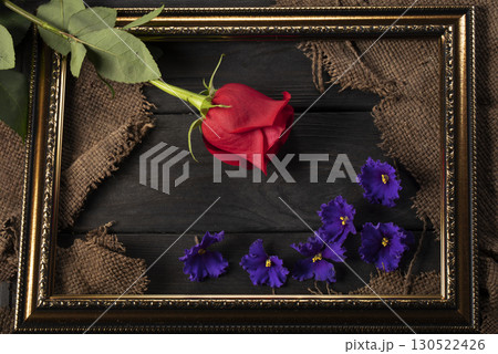 In a beautiful frame, pieces of canvas, a red rose and violets on a wooden background. Decor and decoration. In a beautiful frame, pieces of canvas, a red rose and violets on a wooden background. Decor and decoration. 130522426
