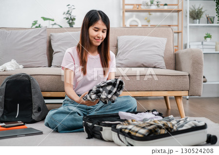 Smiling woman organizing clothes in a suitcase, preparing for a trip in a comfortable living room setting. 130524208