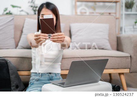 Woman holding a passport, sitting on a suitcase with a laptop, ready for travel in a modern living room. 130524237