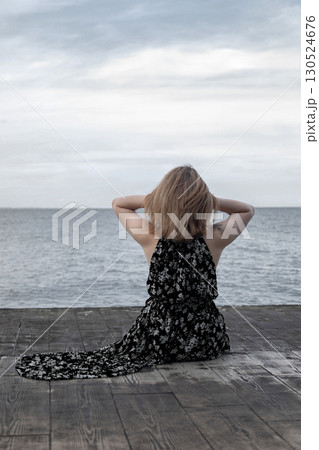 Woman in dress sitting on wooden pier by the sea, back view. Concept of relaxation, solitude, freedom, summer vacation and peaceful lifestyle Woman in dress sitting on wooden pier by the sea, back view. Concept of relaxation, solitude, freedom, summer vacation and peaceful lifestyle 130524676