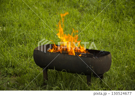 Bright fire burning in barbecue grill on green grass. Concept of outdoor cooking, picnic preparation, camping, summer lifestyle and fire element Bright fire burning in barbecue grill on green grass. Concept of outdoor cooking, picnic preparation, camping, summer lifestyle and fire element 130524677