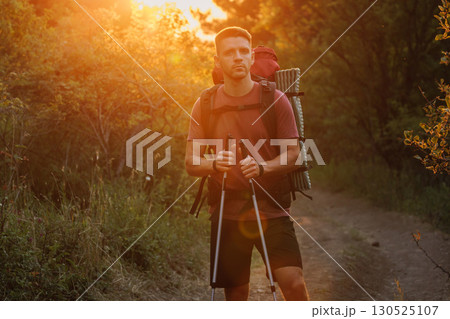 Handsome man hiker with backpack and poles walking through forest with warm sunset. Handsome man hiker with backpack and poles walking through forest with warm sunset. 130525107