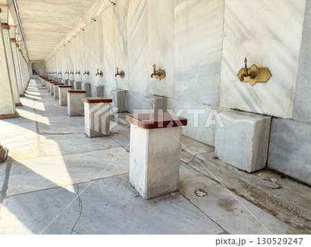 Row of brass water taps with stone seats arranged for ritual ablution. Religion, culture, and daily practice of tradition in architectural setting. 130529247
