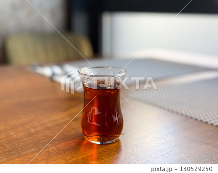 Glass of hot turkish tea served on a wooden table in a dining setting. Hospitality, relaxation, and traditional beverage culture in everyday lifestyle and social moment. Glass of hot turkish tea served on a wooden table in a dining setting. Hospitality, relaxation, and traditional beverage culture in everyday lifestyle and social moment. 130529250