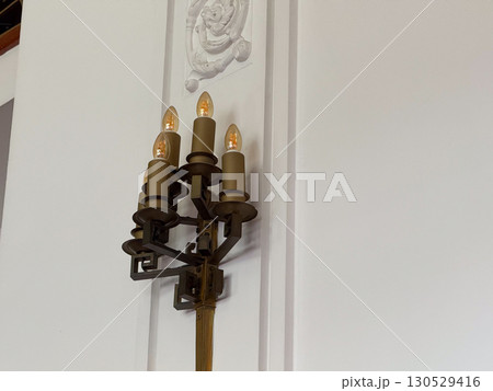 Bronze wall sconce with four bulbs designed in candle style on a white wall. Architectural detail, vintage lighting, and heritage decor in historic interior design. 130529416