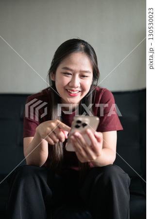 Young Thai woman sitting on sofa using smartphone at home, lifestyle portrait Young Thai woman sitting on sofa using smartphone at home, lifestyle portrait 130530023