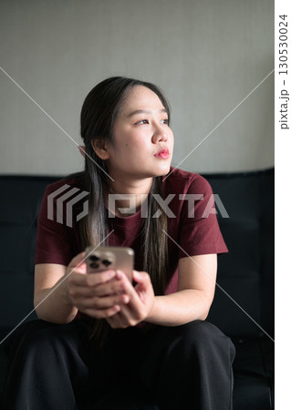 Young Thai woman sitting on sofa using smartphone at home, lifestyle portrait 130530024