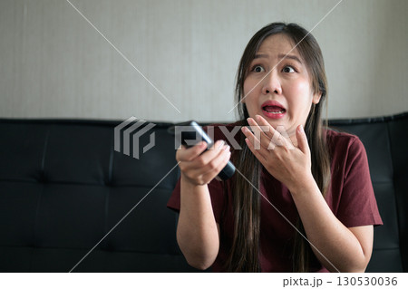 Thai woman sitting on sofa watching television at home looking stressed and scared 130530036