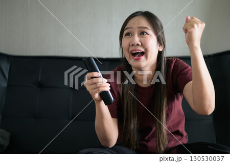 Thai woman sitting on sofa watching television at home looking happy and excited 130530037