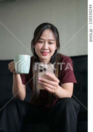 Young Thai woman sitting on sofa using smartphone at home and drinking coffee 130530244
