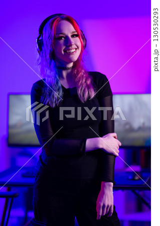 Colorfully lit young female gamer with headphones smiling confidently 130530293