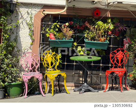 Brightly painted chairs and a green table placed outdoors near a window with plants. Decoration, leisure, and lifestyle in urban cafe atmosphere. Brightly painted chairs and a green table placed outdoors near a window with plants. Decoration, leisure, and lifestyle in urban cafe atmosphere. 130530692
