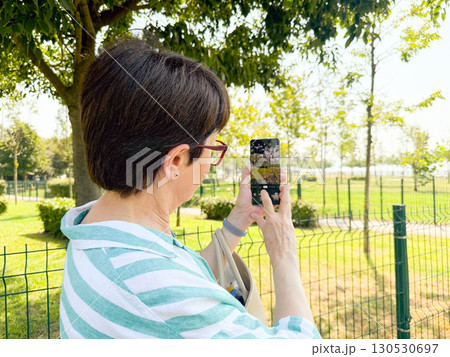 Woman taking a photo outdoors with a smartphone in her hands. Daily life, urban leisure, and personal perspective in an open park environment. Woman taking a photo outdoors with a smartphone in her hands. Daily life, urban leisure, and personal perspective in an open park environment. 130530697