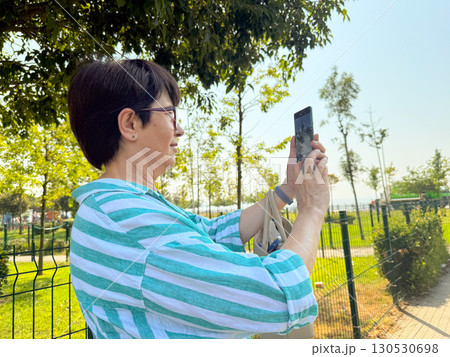 Woman taking a photo outdoors with a smartphone in her hands. Daily life, urban leisure, and personal perspective in an open park environment. 130530698