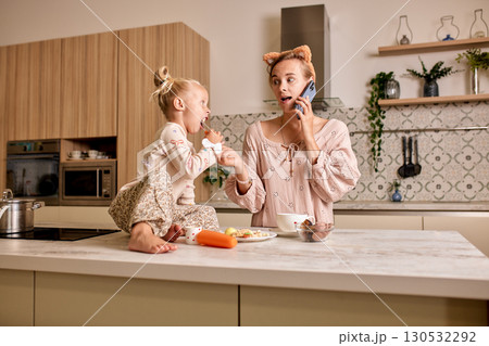 Mother multitasking on phone while bonding with daughter in modern kitchen 130532292