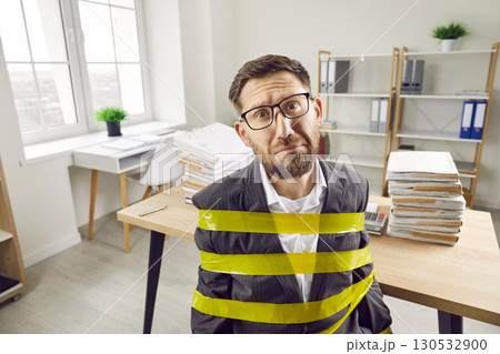 Funny office slave, wrapped with adhesive tape, sitting by desk with load of paperwork 130532900