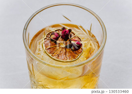 Close-up of cocktail with large ice cube, dried citrus slice and rosebuds in a crystal glass 130533460