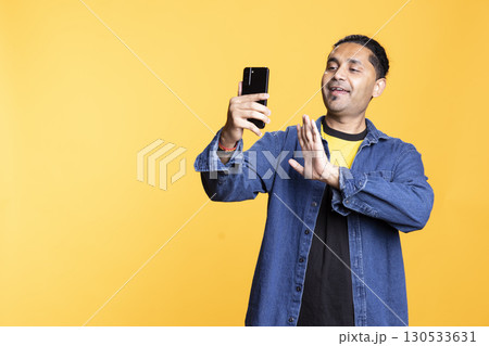 Relaxed man having entertaining conversation with a mate over online videocall on smartphone. Carefree indian guy discussing with friends on teleconference, studio background. 130533631